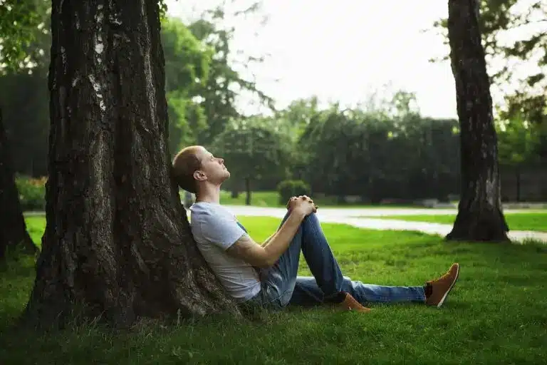 A man leans quietly against a tree, eyes closed, grounded in stillness. The gentle light filtering through the leaves evokes a sense of calm and reflection. This image captures the essence of the podcast episode “How to Create Safety After Childhood Trauma”—a journey of returning to inner calm, reconnecting with the body, and discovering what safety can feel like after trauma. Just as the tree offers steady support, healing asks us to root ourselves in care, presence, and patience.