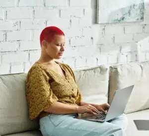 A woman of colour sitting comfortably on a sofa with a laptop on her lap, smiling warmly. She represents a client engaged in trauma-informed healing, connecting online with an experienced and skilled trauma counsellor for personalized support and growth.