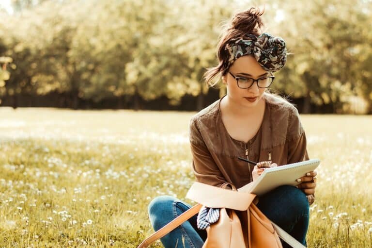 A woman sitting outside under a tree, journaling in a notebook, reflecting quietly — illustrating the concept of what is retraumatization and ways to process trauma safely.