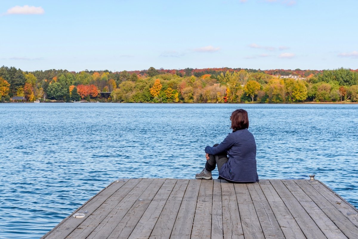 Woman representing adults with childhood trauma sitting on a wooden pier overlooking a calm lake on a clear autumn day, reflecting on healing and personal growth.