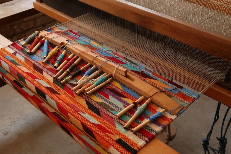 Traditional wooden loom with an unfinished, colorful woven carpet and vibrant threads draped across it, symbolizing how different strands of healing approaches come together in one tapestry, reflecting the integrative nature of IFS or EMDR for complex trauma.